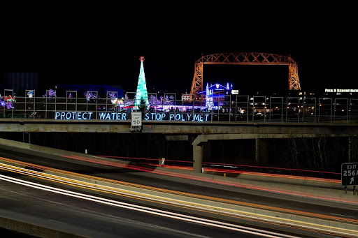 people standing on a bridge at night with a lit sign that says protect water stop polymet