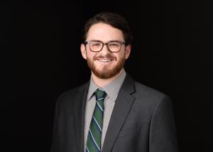 a white man with a beard, smiling, standing in a gray suit and tie