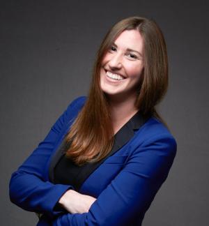 Photo of Paige Stradley. A professional headshot photograph of white woman with long brown hair, smiling with arms crossed, wearing a blue suit.