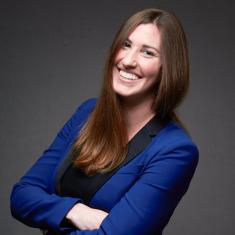 Photo of Paige Stradley. A professional headshot photograph of white woman with long brown hair, smiling with arms crossed, wearing a blue suit.