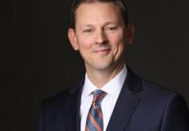 a white man with short hair smiling in a suit on a dark background